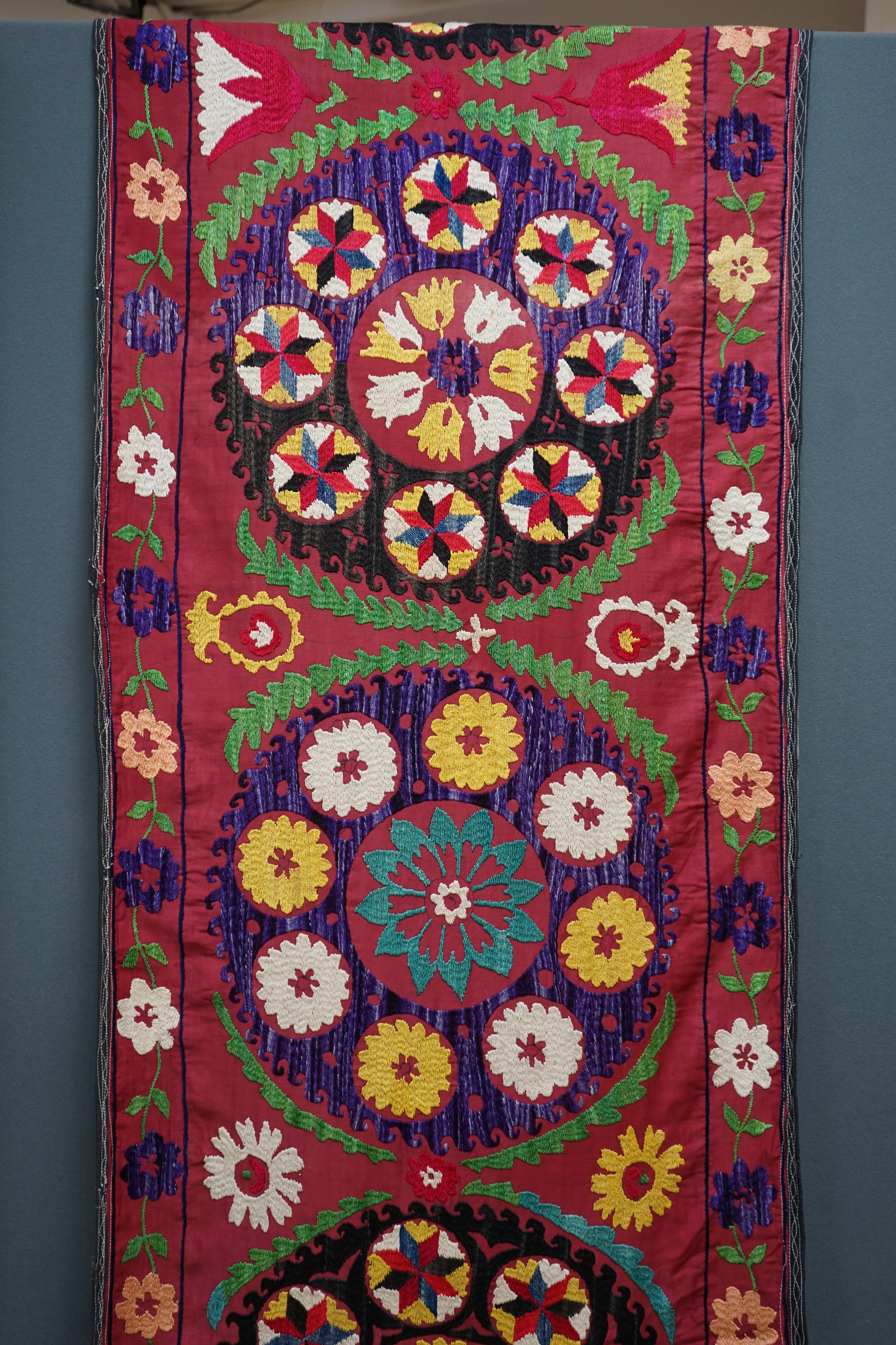 A 20th century Suzani wall hanging, possibly Uzbekistan, embroidered in bright polychrome silk threads into large symbols representing happiness, health and luck, on a burgundy cotton background, with a fine muslin backi